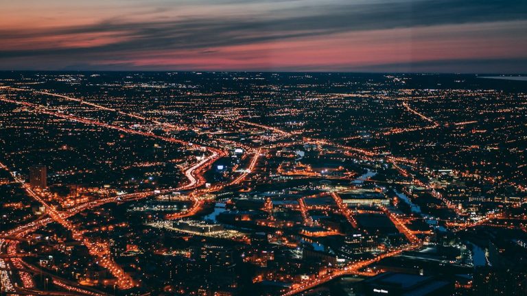 Aerial night view of Boston, MA with glowing city streets and highways stretching to the horizon at dusk.